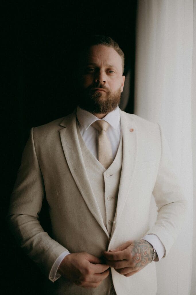 Groom by window on wedding day in tan tuxedo